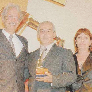 Na imagem, há a fotografia de três pessoas, sendo dois homens usando traje social completo e uma mulher utilizando roupa preta social. No canto esquerdo, há um homem de cabelos brancos sorrindo para a câmera e, abraçado a ele, há outro homem segurando um troféu nas mãos. Do lado direito, a mulher sorri discretamente para a câmera.