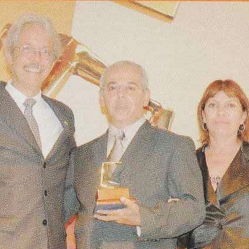 Na imagem, há a fotografia de três pessoas, sendo dois homens usando traje social completo e uma mulher utilizando roupa preta social. No canto esquerdo, há um homem de cabelos brancos sorrindo para a câmera e, abraçado a ele, há outro homem segurando um troféu nas mãos. Do lado direito, a mulher sorri discretamente para a câmera.
