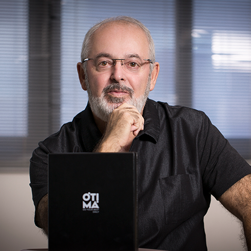 Na imagem, há a fotografia de um homem de meia-idade, de óculos e com cabelos e barba brancos. Ele está sentado e olha diretamente para a câmera, apoiando o cotovelo esquerdo no tampo de uma mesa onde há um caderno com o logo da Ótima Gráfica em destaque.
