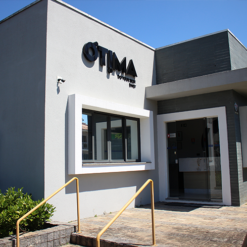 Na imagem, há a fotografia da entrada da sede da Ótima Gráfica, um prédio de dois andares com a pintura na cor cinza e o logo da Ótima em destaque, na cor preta.